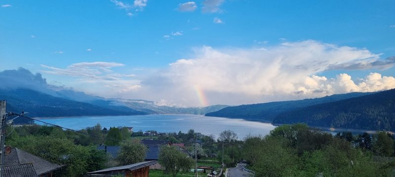 Panoramă largă asupra Lacului Bicaz, cu apa albastră întinsă între dealuri împădurite și nori pe cer